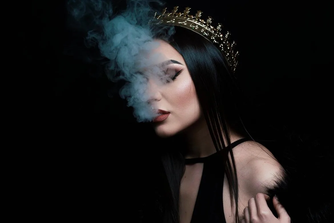 A woman wearing a jeweled crown as smoke drifts across her face against a black background.