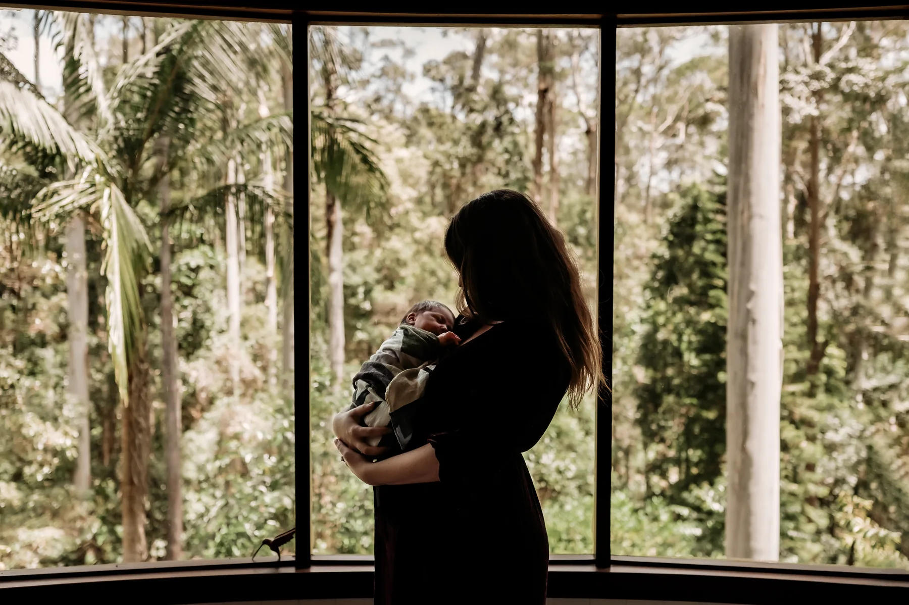 A mother holding her newborn in silhouette beside a large window.