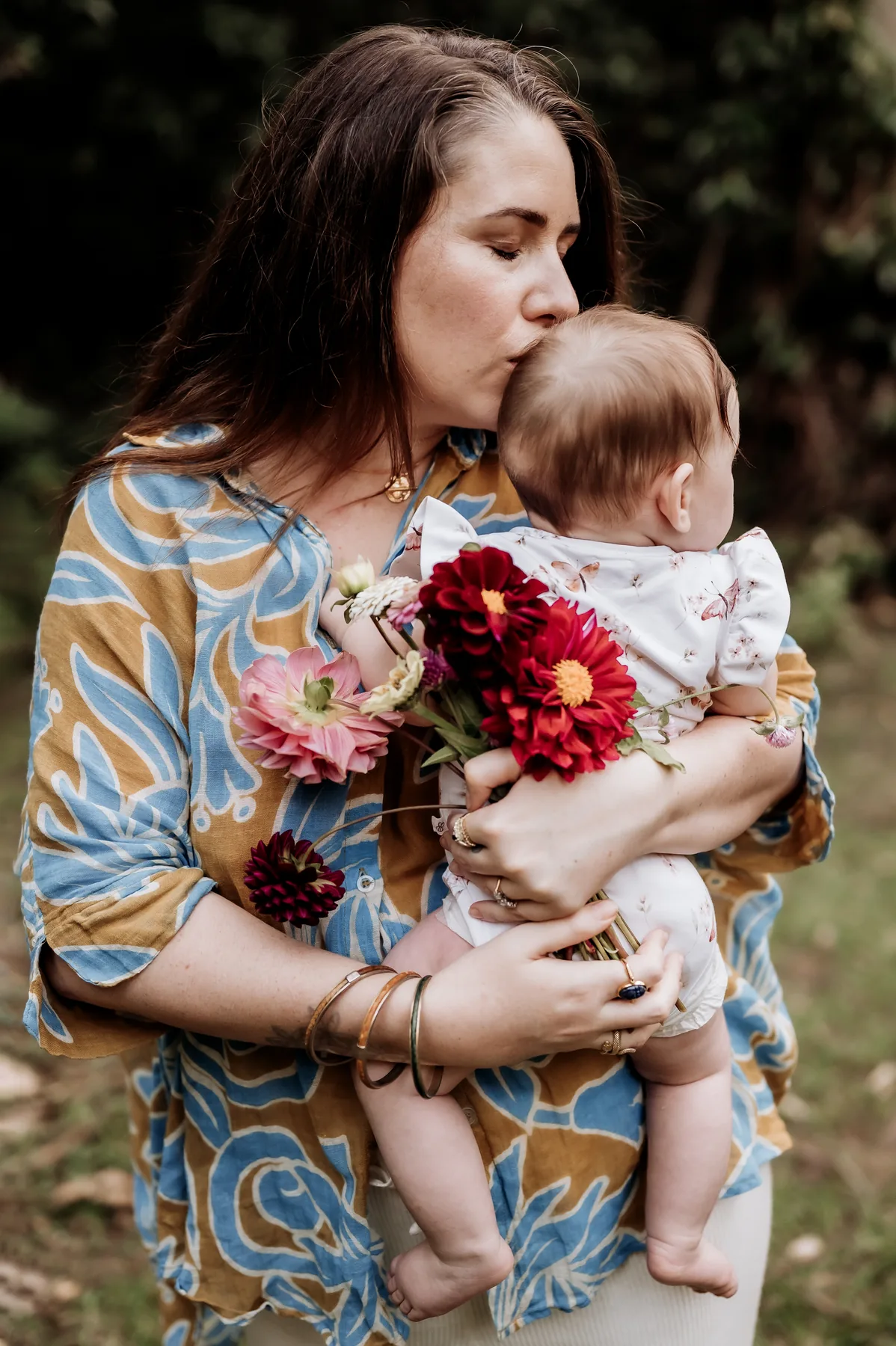 A mother kissing her baby while holding a bouquet of flowers outdoors.